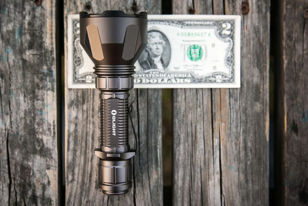 A black flashlight is placed on a weathered wooden surface, positioned above a two-dollar bill for size comparison.