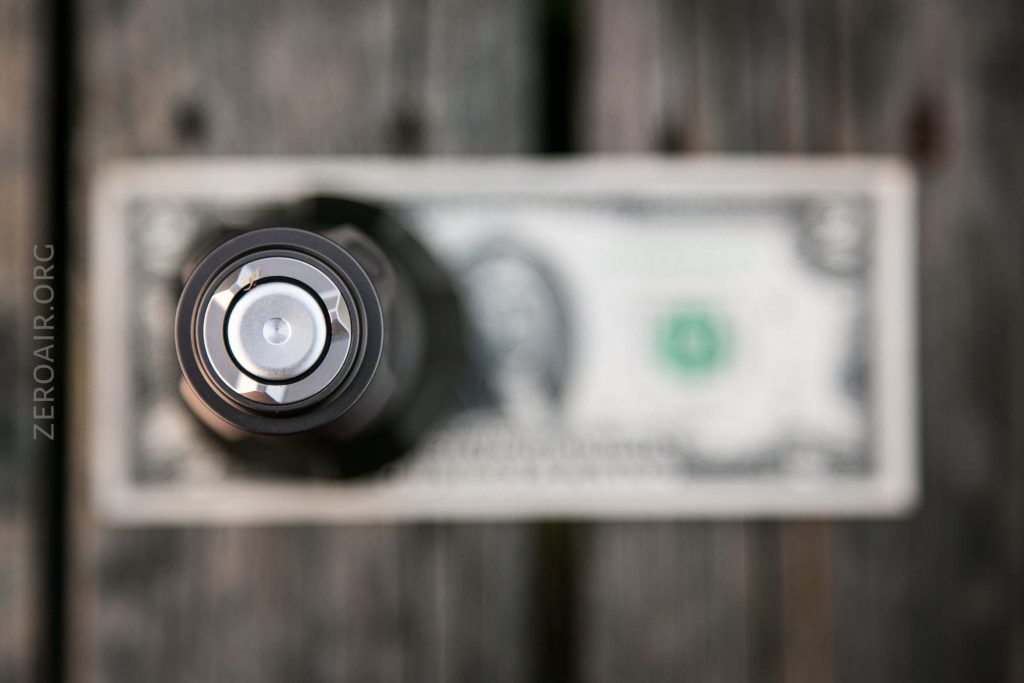 A close-up, top-down view of a metallic cylinder, possibly a flashlight, with a U.S. one dollar bill blurred in the background on a wooden surface. The cylinder is centered and in sharp focus.