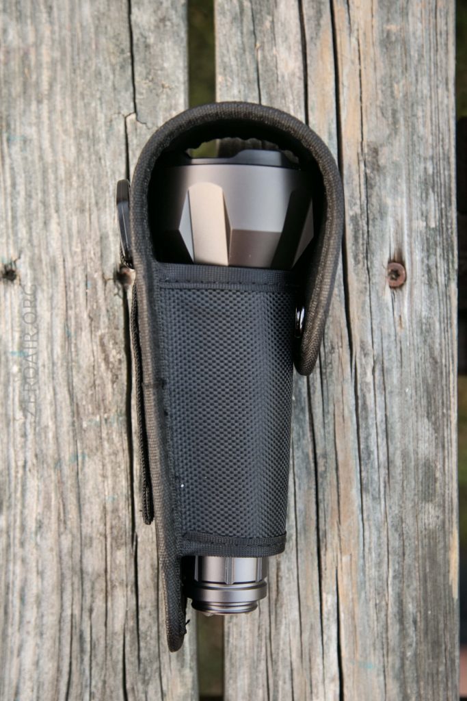 A metallic flashlight partially enclosed in a black nylon holster, secured to weathered wooden planks with visible grain and cracks.