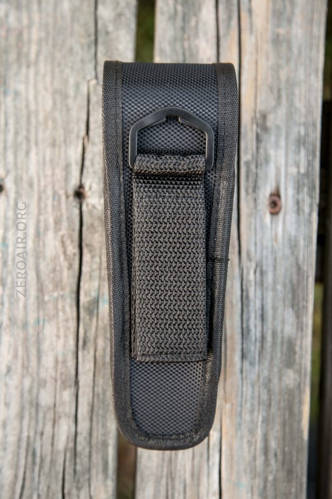 A black nylon pouch with a textured strap and metal D-ring is attached to a weathered wooden surface. The pouch appears to be designed for carrying tools or small equipment.