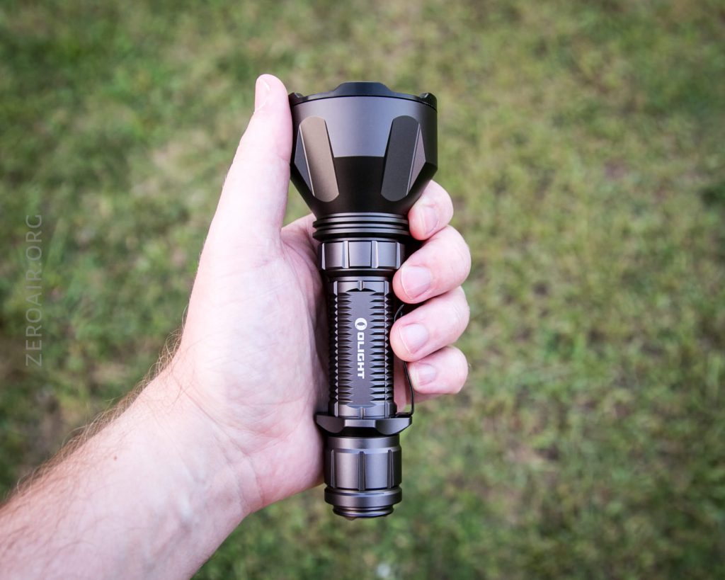 A hand holds a black, metallic flashlight with a ridged grip, wide head, and the brand name “Olight” visible on the body. The background is out-of-focus green grass.