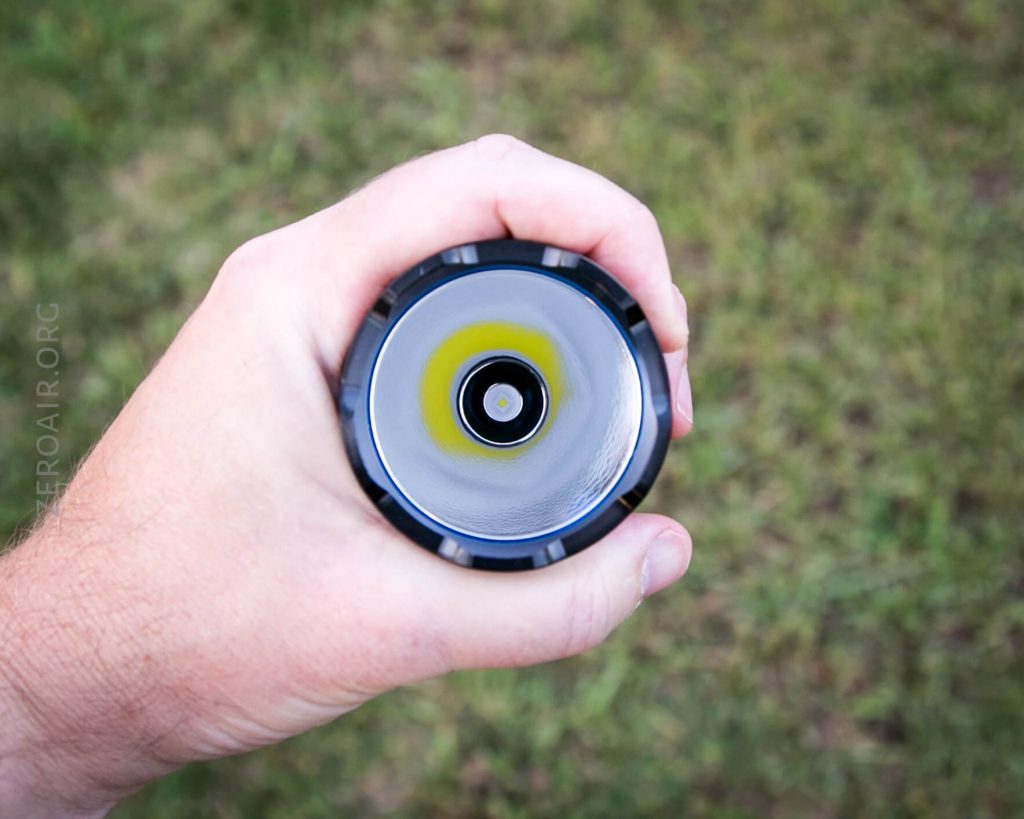 A hand holds a black flashlight, viewed from above, showing the lens and central LED bulb. The background is grassy and slightly out of focus.