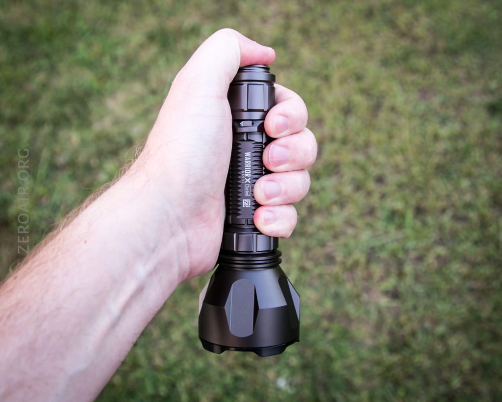 A hand holding a large black flashlight outdoors, with grass visible in the background.