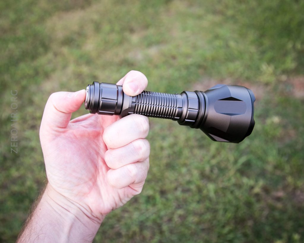 A hand holding a black flashlight outdoors, with grass visible in the background. The flashlight has a ribbed grip and a wide head.
