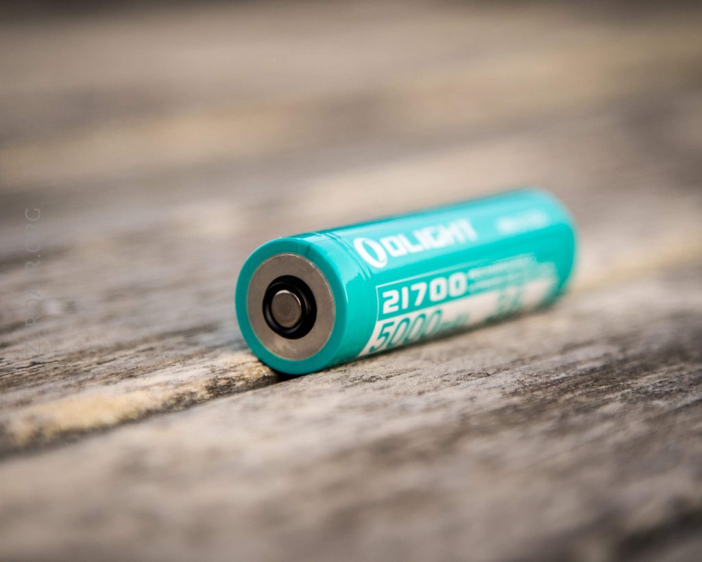 A teal Olight 21700 rechargeable lithium-ion battery with a capacity of 5000mAh lies horizontally on a weathered wooden surface.