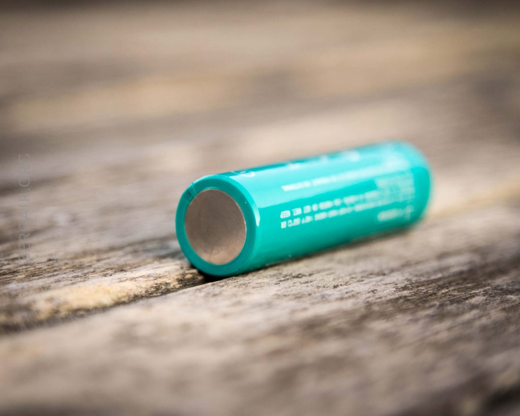 A close-up of a teal cylindrical battery lying horizontally on a rough wooden surface, with the flat, metal end facing the camera. The batterys text and markings are partially visible.