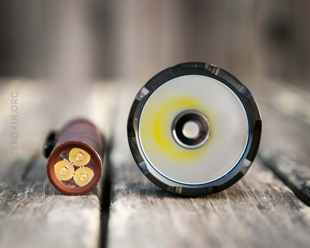 A close-up of a flashlight head showing a yellow LED in the center, next to a cylindrical battery with three LED components, both placed on a wooden surface.