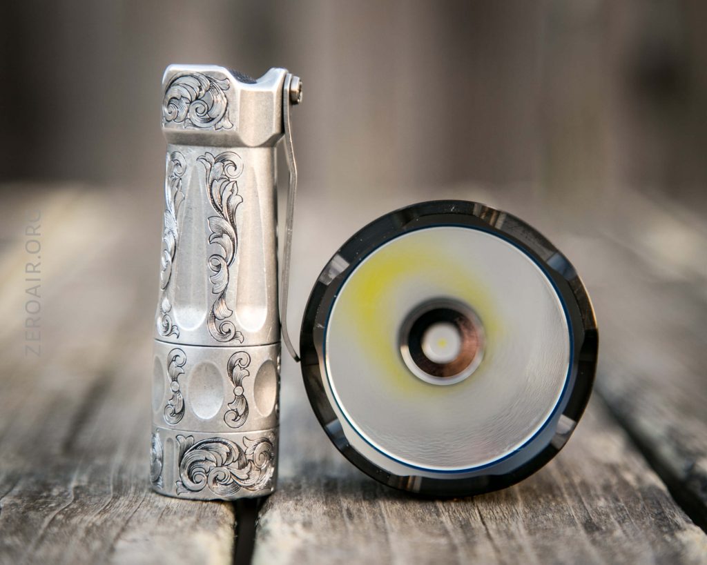 A metallic flashlight with ornate engraved patterns stands upright next to its open lens, both resting on a weathered wooden surface. The inside reflector and LED bulb of the flashlight are visible.