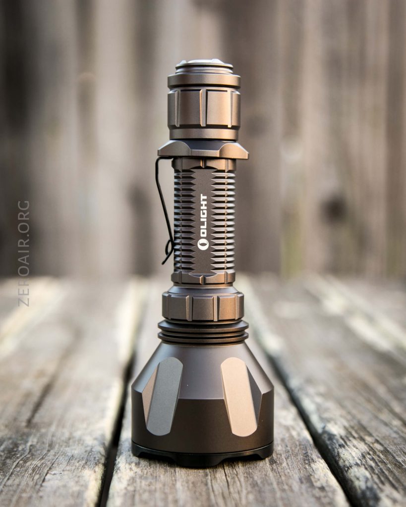 A metallic Olight flashlight stands upright on a wooden surface outdoors, with a blurred wooden fence in the background. The flashlight has a ribbed midsection and angular base.