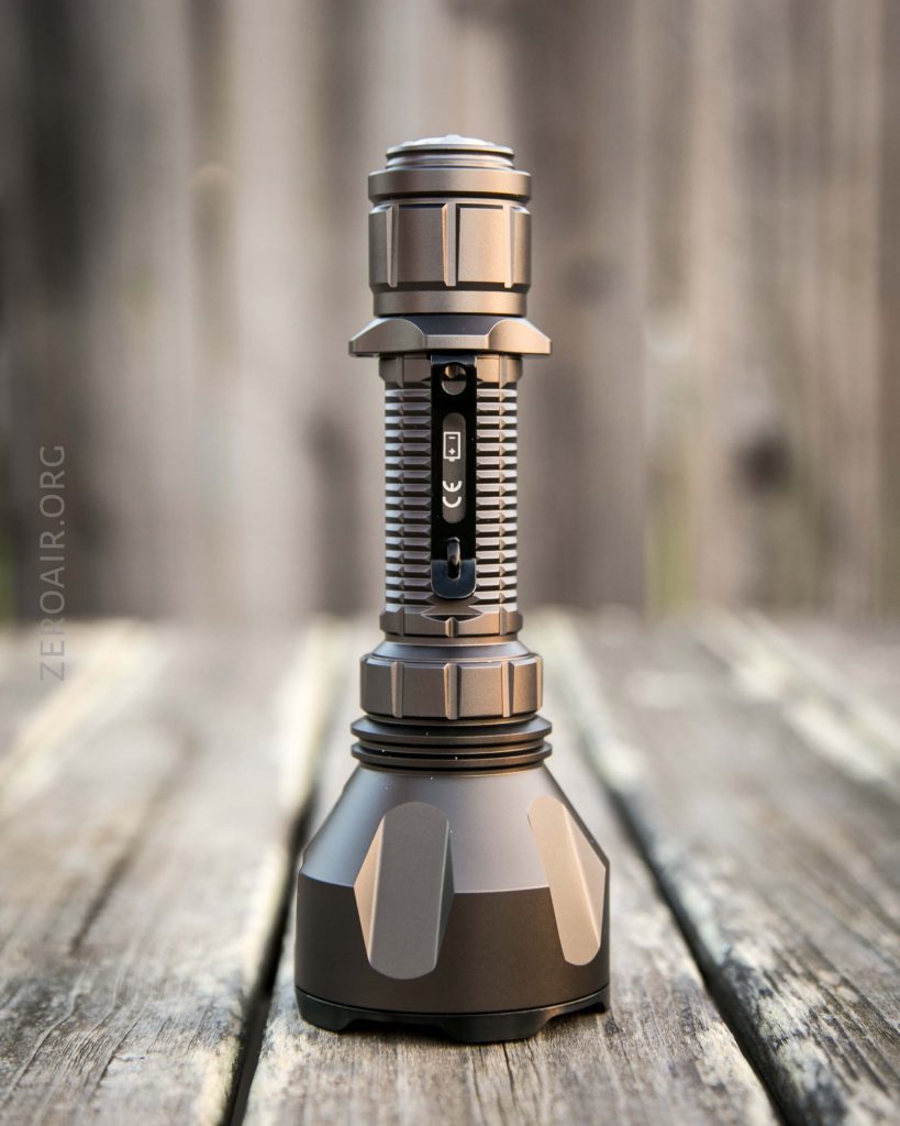 A metallic flashlight with a ribbed grip and a wide base stands upright on a wooden surface, with a blurred wooden fence in the background. The flashlight’s branding and markings are visible on the side.