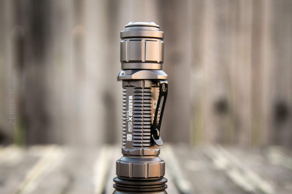 A metallic flashlight with a textured grip and a side clip is standing upright on a wooden surface, with a blurred wooden background.