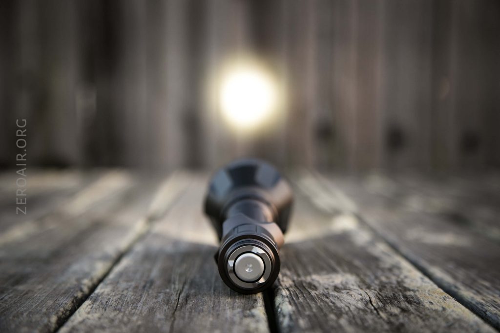 A black flashlight lies on a weathered wooden surface, shining a bright beam of light forward. The image is focused on the flashlights button, with the background and light appearing blurred.