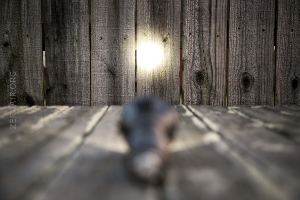 A wooden fence with light shining through a hole in one plank. The foreground is blurred, showing an indistinct dark object on a weathered wood surface.