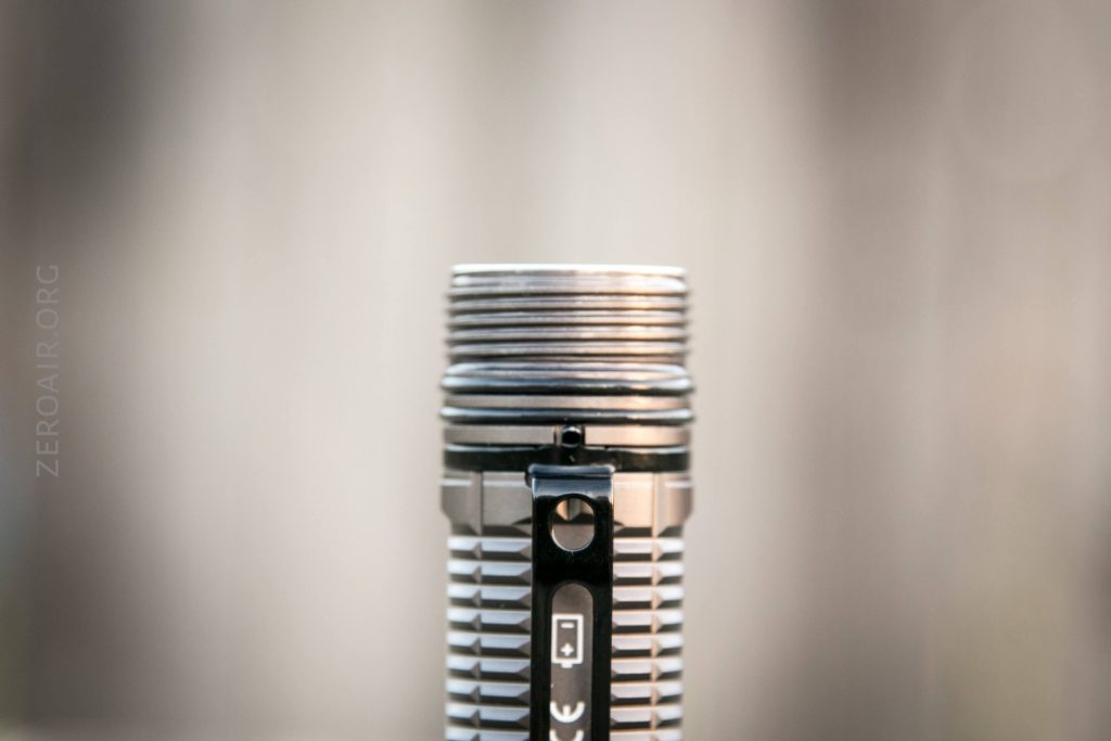 A close-up of the top portion of a silver flashlight with a black clip attached, shown against a blurred neutral background. The battery symbol and some text are visible on the clip.