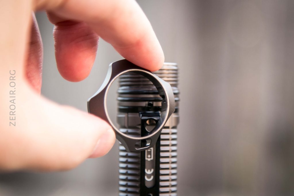 A hand holds a metal ring-shaped object above the top of a cylindrical, metallic, ribbed device. The background is blurred and ZEROAIR.ORG is visible on the left side of the image.