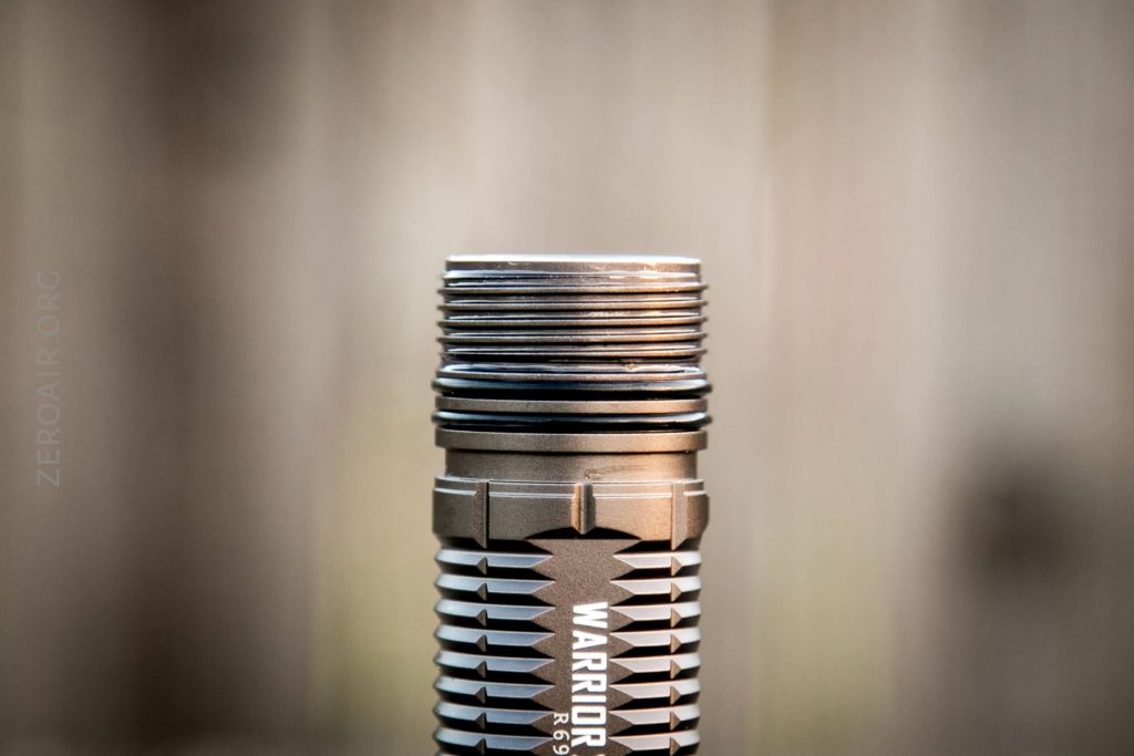 A close-up of the threaded top section of a metallic flashlight with WARRIOR and textured grip visible, set against a blurred neutral background.