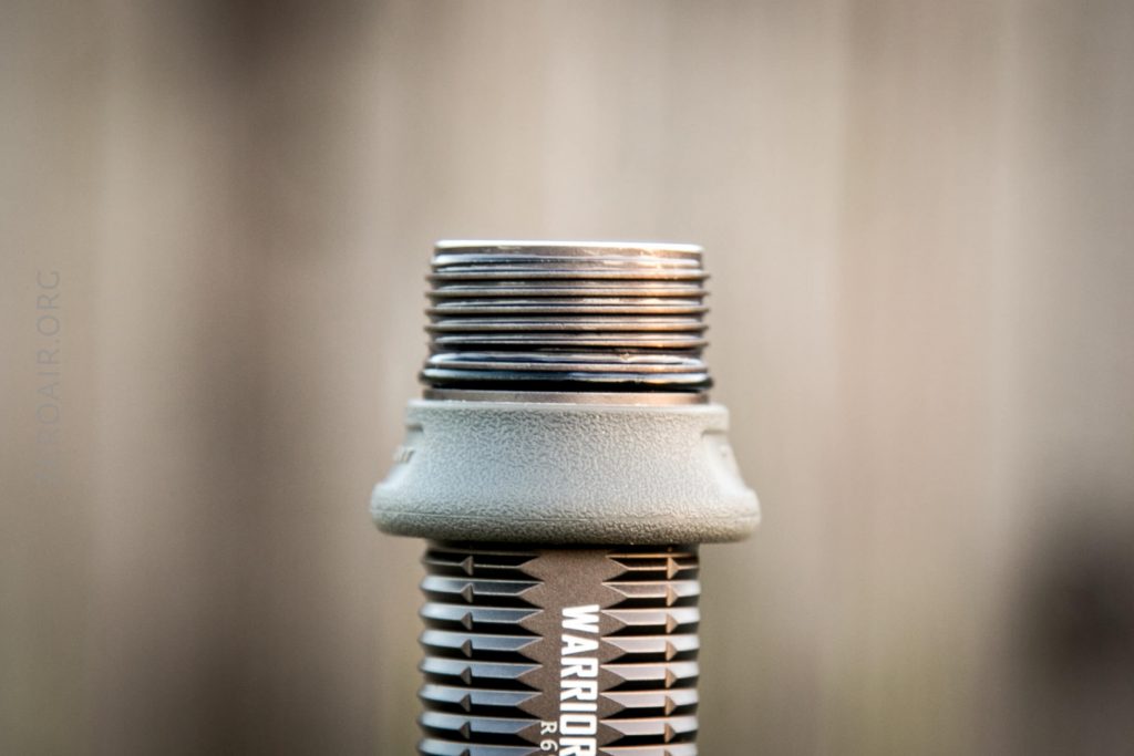 A close-up view of the threaded end of a cylindrical metal object labeled WARRIOR, with visible grooves and a textured grip. The background is blurred and neutral in color.