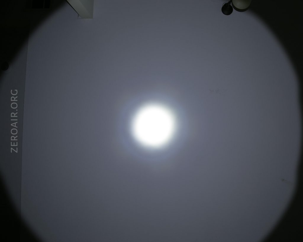 A bright flashlight beam creates a circular spot on a plain wall, with a strong central hotspot and a dimmer outer ring. The text ZEROAIR.ORG is visible on the left side of the image.