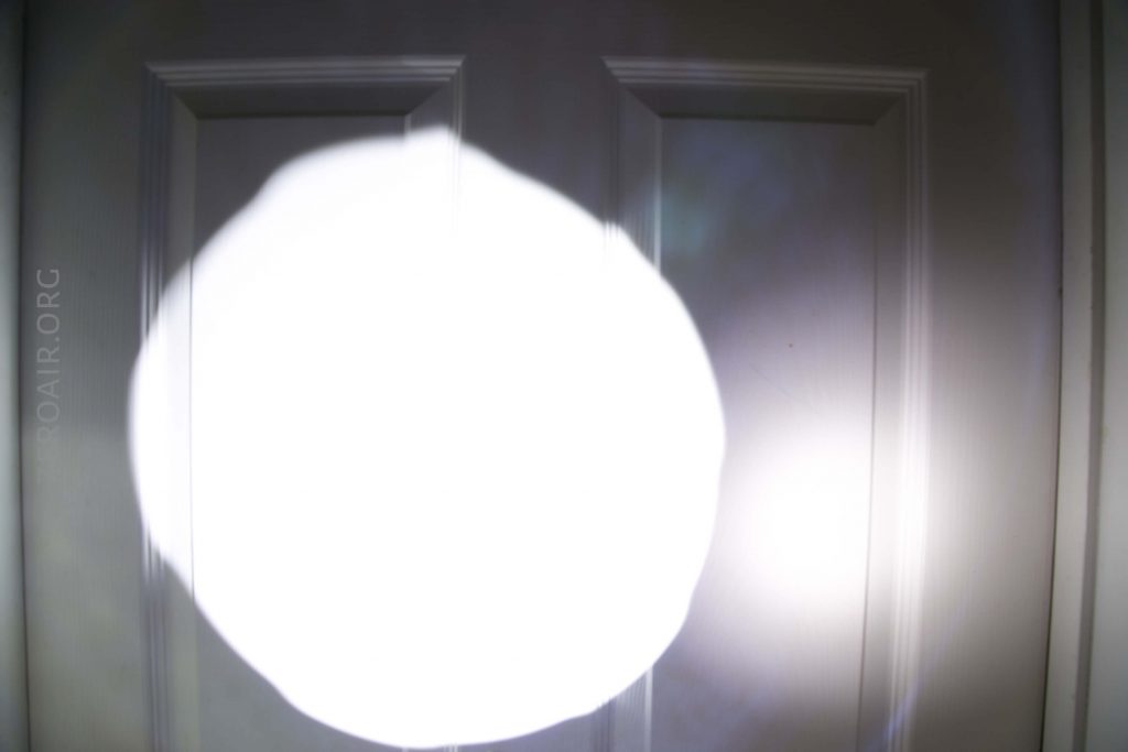 A bright white circular light is projected onto a white paneled door, creating a strong contrast between the illuminated and surrounding areas. The door has vertical rectangular panels and is mostly obscured by the light.