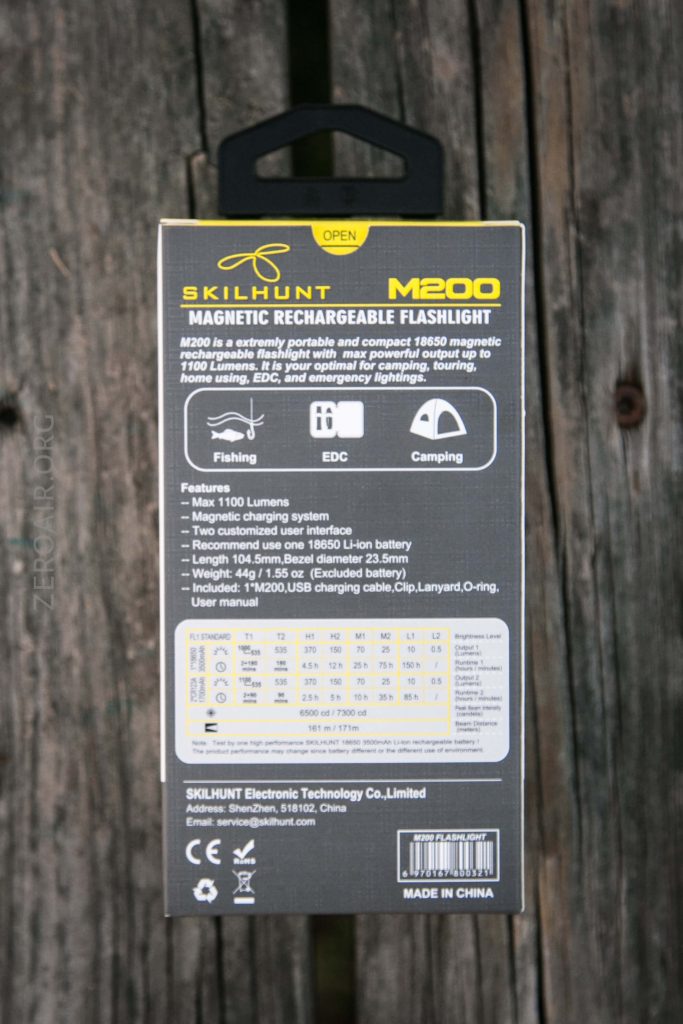 A Skilhunt M200 magnetic rechargeable flashlight box stands upright on a wooden surface, showing product details, features, specifications, icons for fishing, EDC, and camping, and a performance chart on the back.