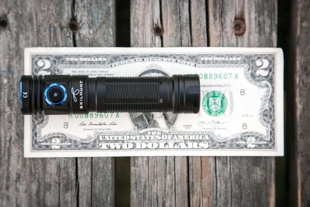 A black Skilhunt flashlight is placed on top of a two-dollar bill, both resting on weathered wooden planks.
