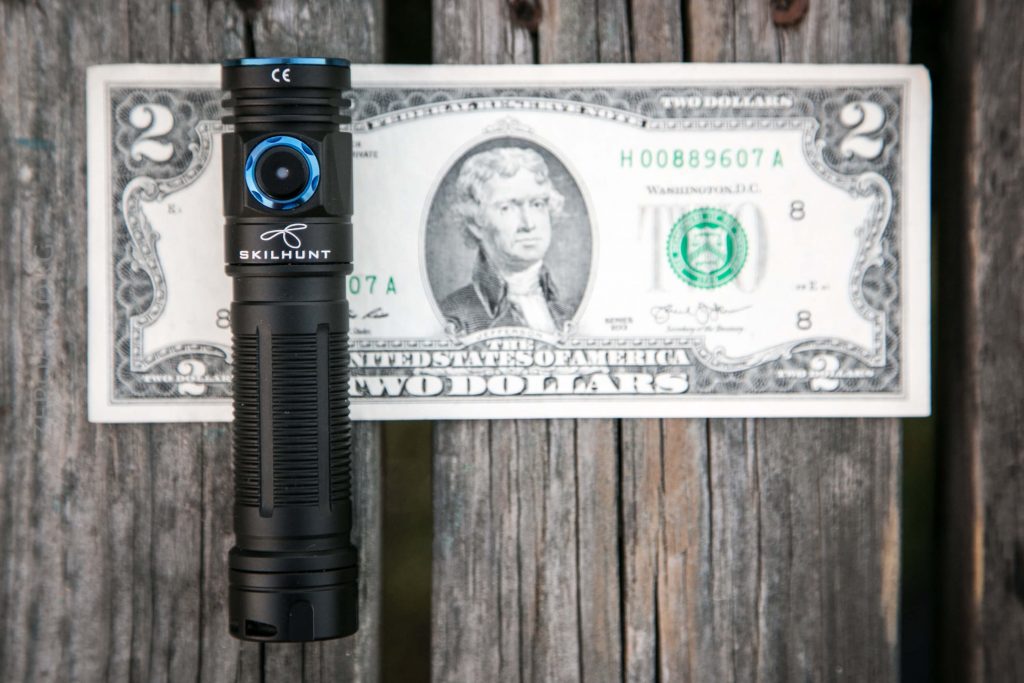 A black Skilhunt flashlight is standing upright in front of a U.S. two-dollar bill, which is placed on a weathered wooden surface. The flashlight partially covers the bills portrait of Thomas Jefferson.