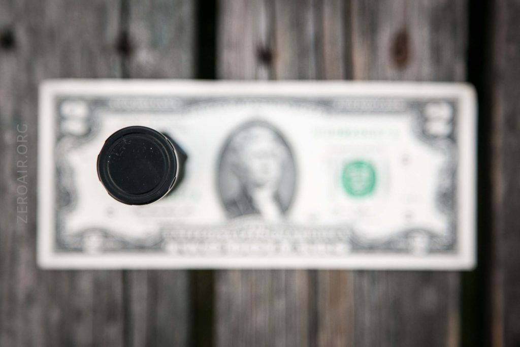 A black cylindrical object viewed from above is centered on top of a horizontally placed two-dollar bill, which is lying on a wooden surface. The bill is out of focus.