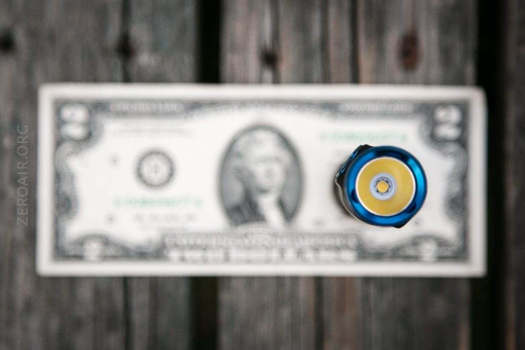 A two-dollar bill lies flat on a wooden surface, with a round object featuring a yellow center and blue outer ring placed upright in the foreground, partially obscuring the bill.