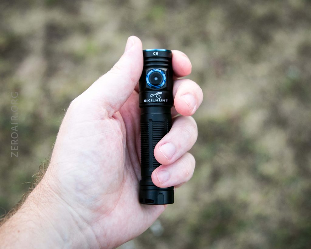 A hand holding a black Skilhunt flashlight with a blue button, against a blurred outdoor background.