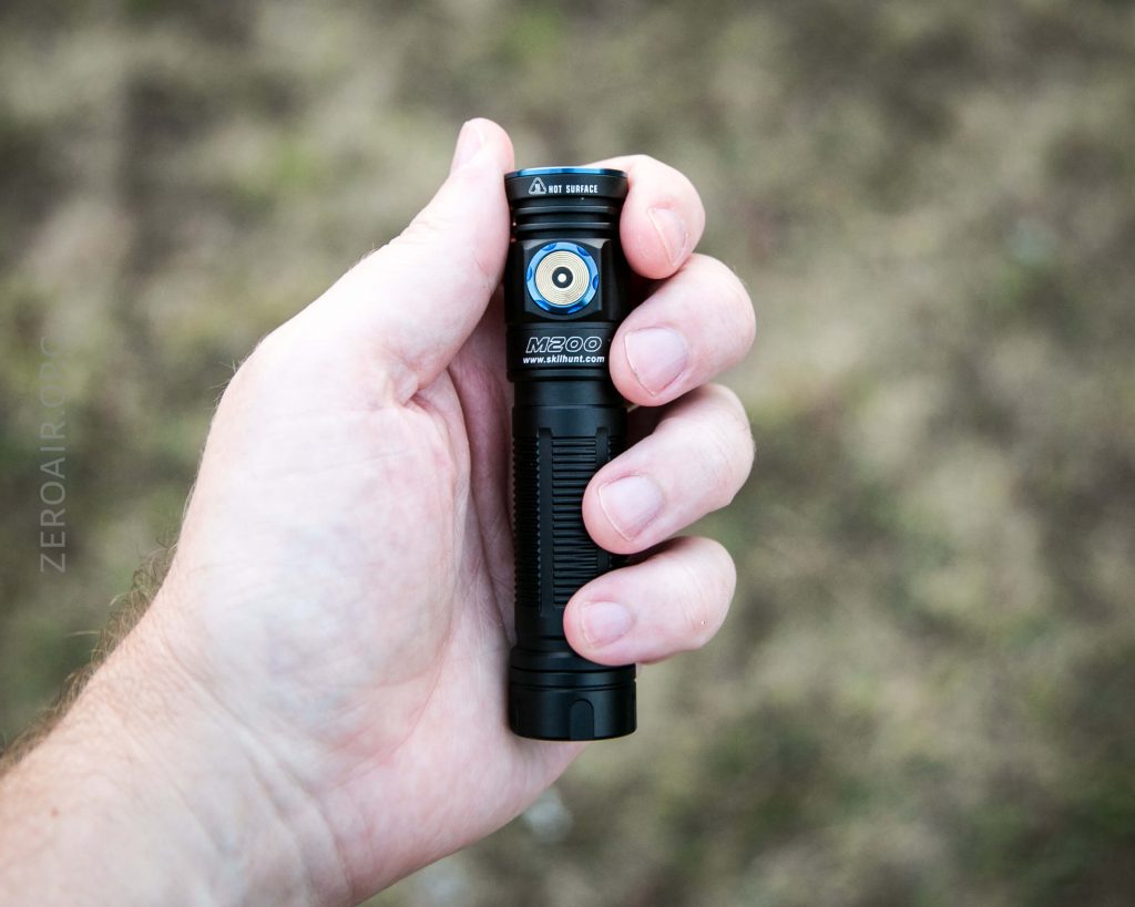 A person holding a black M200 flashlight in their left hand, with an outdoor, blurred grassy background.