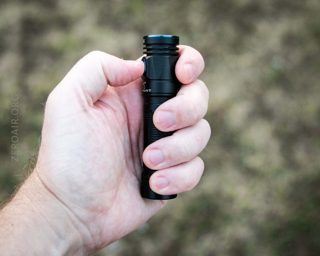 A left hand holding a small black flashlight against a blurred outdoor background.
