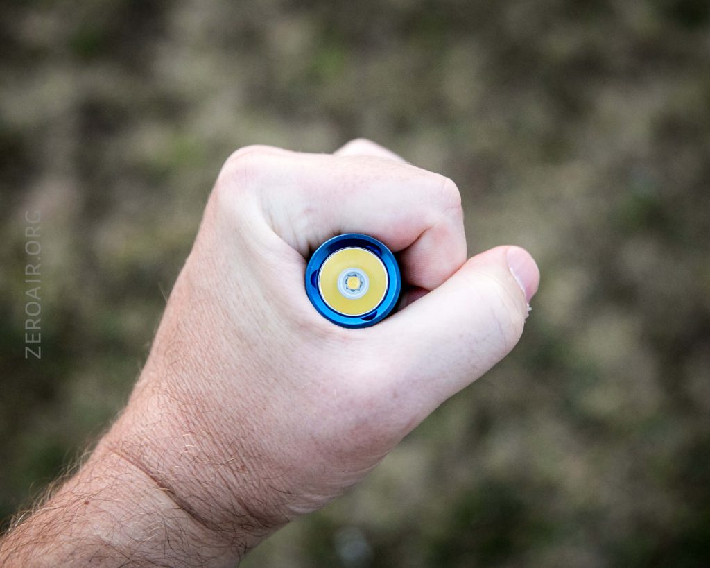 A hand grips a blue and yellow cylindrical object, shown from above against a blurred brown and green background. The object appears to be a battery or similar item.