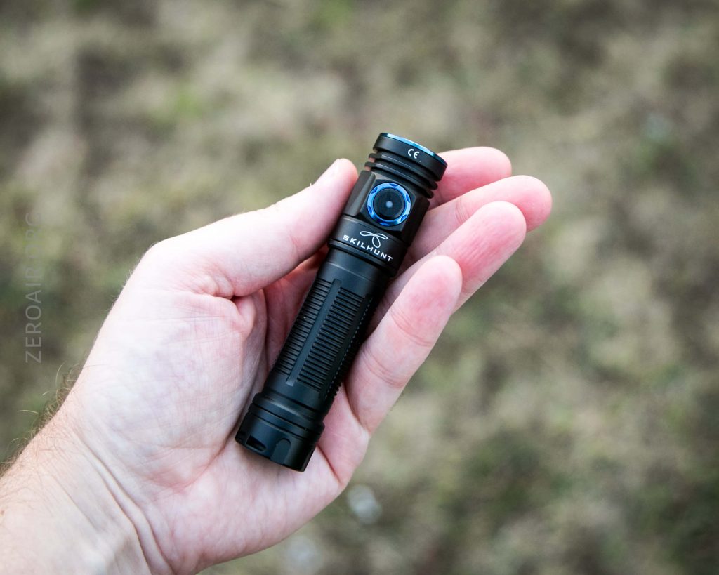 A person holds a black SKILHUNT flashlight in their left hand against a blurred outdoor background of grass and dirt.