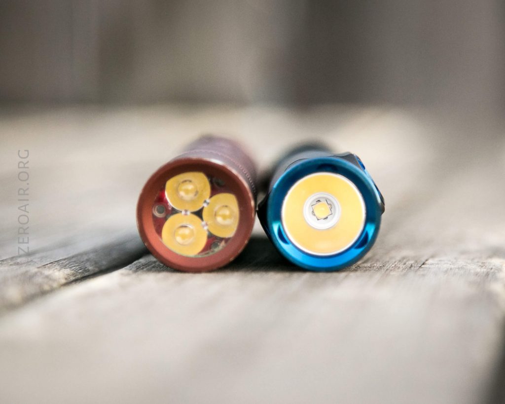 Close-up of two flashlights lying side by side on a wooden surface, showing the LED ends; the left flashlight is red with four LEDs, and the right flashlight is blue with a single LED.