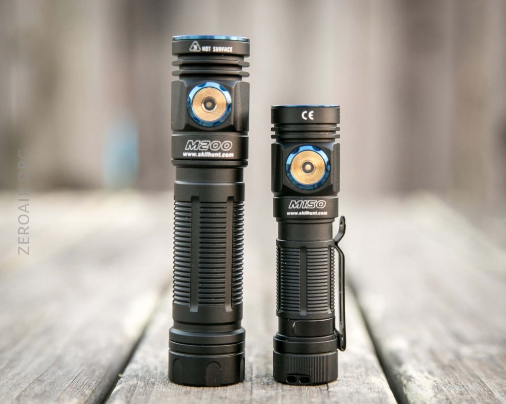 Two black handheld flashlights stand upright on a wooden surface. They differ in height and size, and both have a blue and gold power button on their bodies. Text and model numbers are visible on each flashlight.