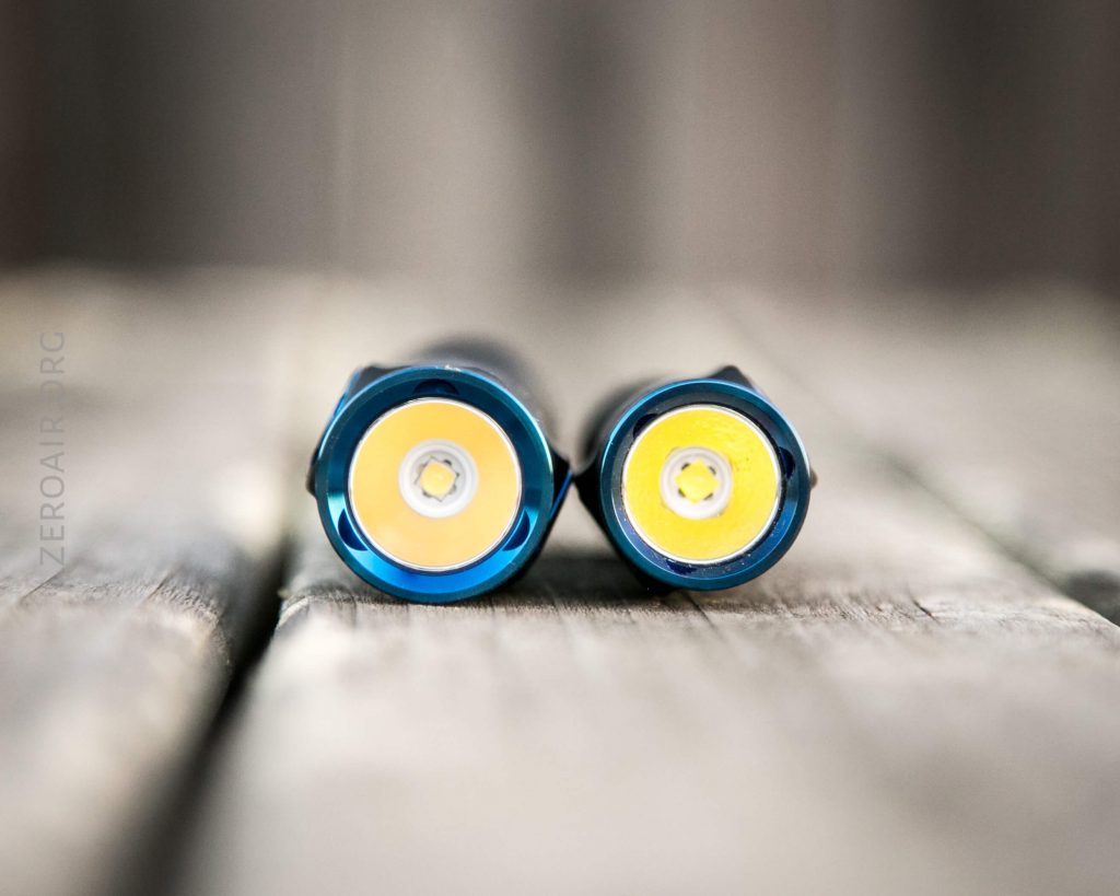 Two small flashlights are shown lying side by side on a wooden surface, viewed from the front with the LED bulbs and reflectors visible. The background is blurred.
