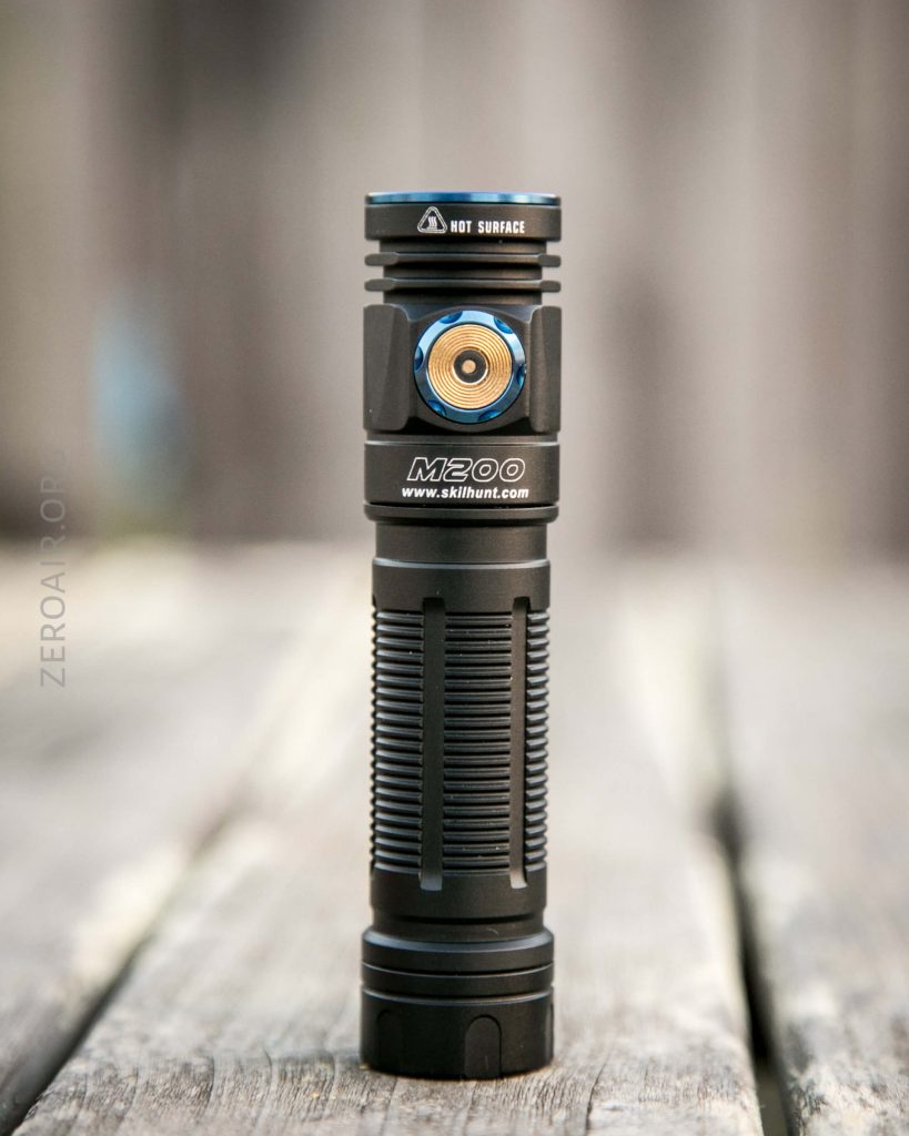 A black Skilhunt M200 flashlight stands upright on a wooden surface with blurred background; the flashlight features a blue-tinted power button and a HOT SURFACE warning label on the head.