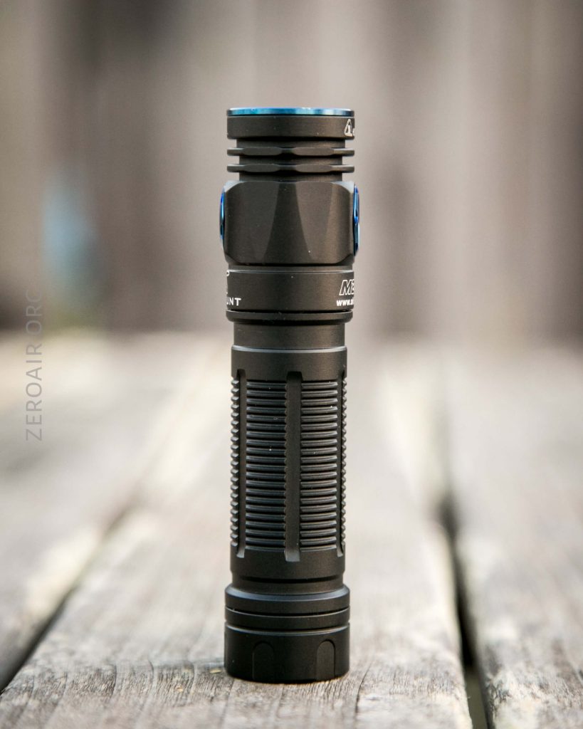A black handheld flashlight with textured grip and blue accents stands upright on a wooden surface with a blurred background.