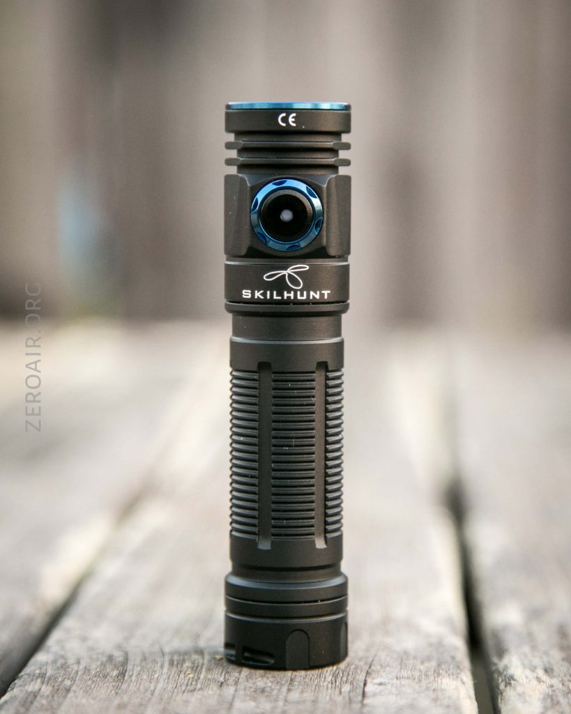 A black Skilhunt flashlight with a blue power button is standing upright on a wooden surface, with a blurred wooden background.