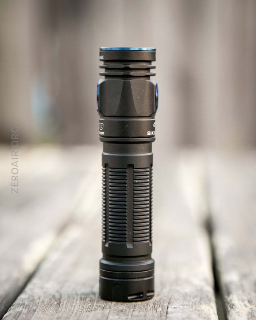 A black handheld flashlight with textured grip and blue accents is standing upright on a wooden surface outdoors. The background is blurred.
