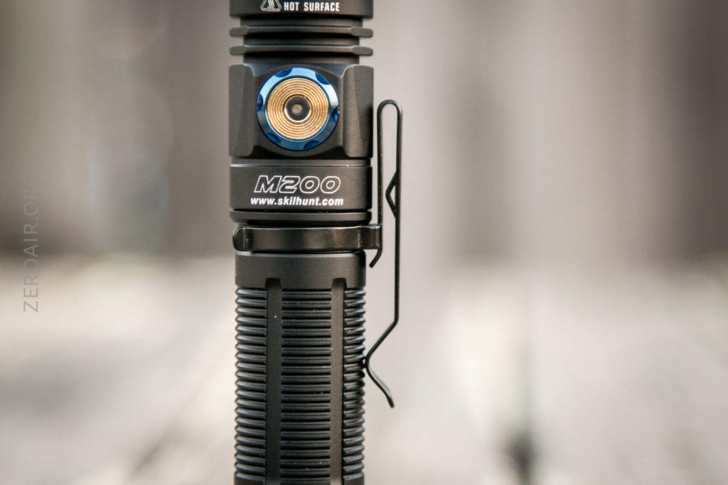 Close-up of a black Skilhunt M200 flashlight, showing its textured grip, metal pocket clip, blue and gold button, and warning label. The background is blurred.
