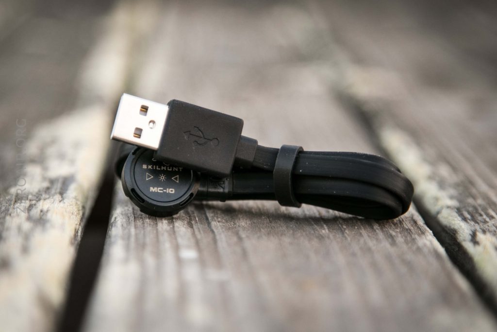 A black USB cable is coiled on a wooden surface. One end shows a USB-A connector, and the other end has a round magnetic charging head labeled SKILHUNT MC-10.