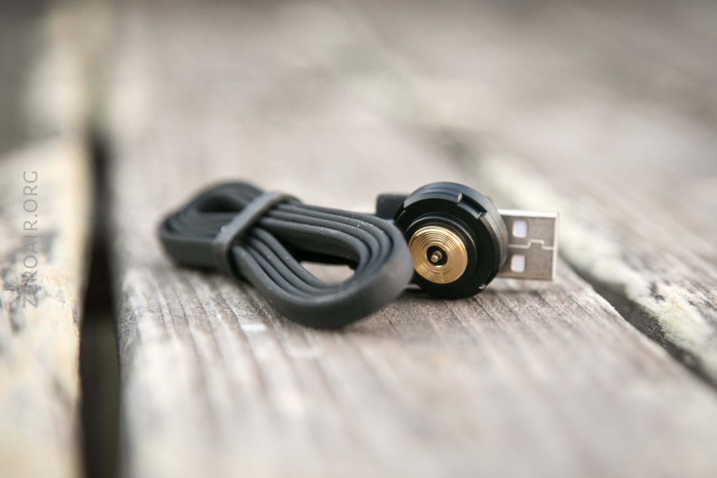 A close-up of a coiled black charging cable with a USB connector on one end and a round, gold and black magnetic connector on the other, lying on a weathered wooden surface.