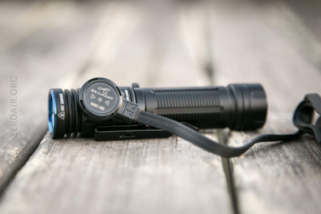 A black flashlight lies on a wooden surface with a close-up of a round magnetic charging cable attached to its side. The background is blurred.