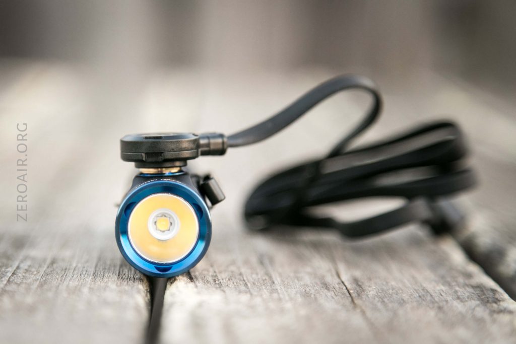 A small blue flashlight with a black cord is resting on a weathered wooden surface. The lens and LED bulb face forward, with the cord coiled and extending out of focus in the background.