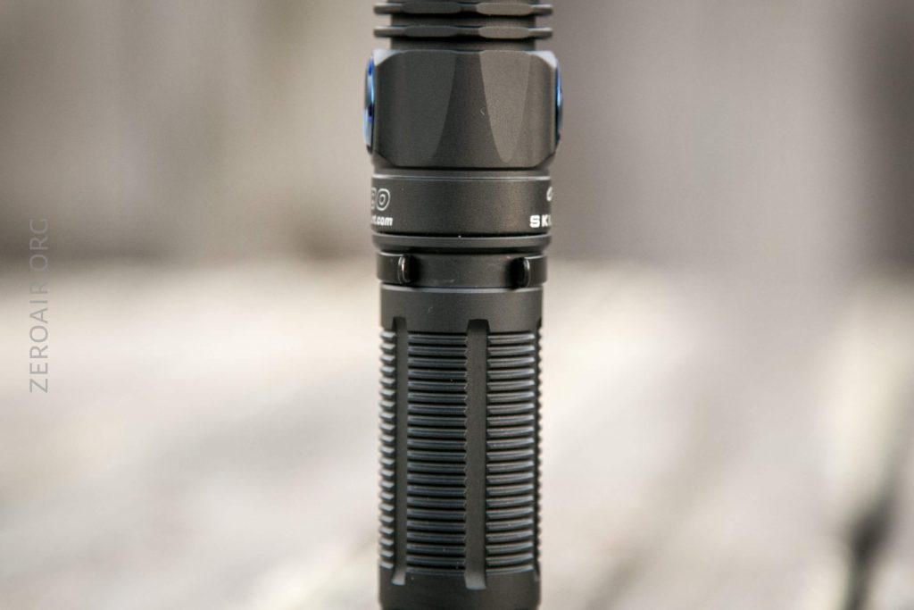 A close-up view of a black, cylindrical flashlight standing upright on a blurred surface, showing textured grip details and part of the manufacturers branding.