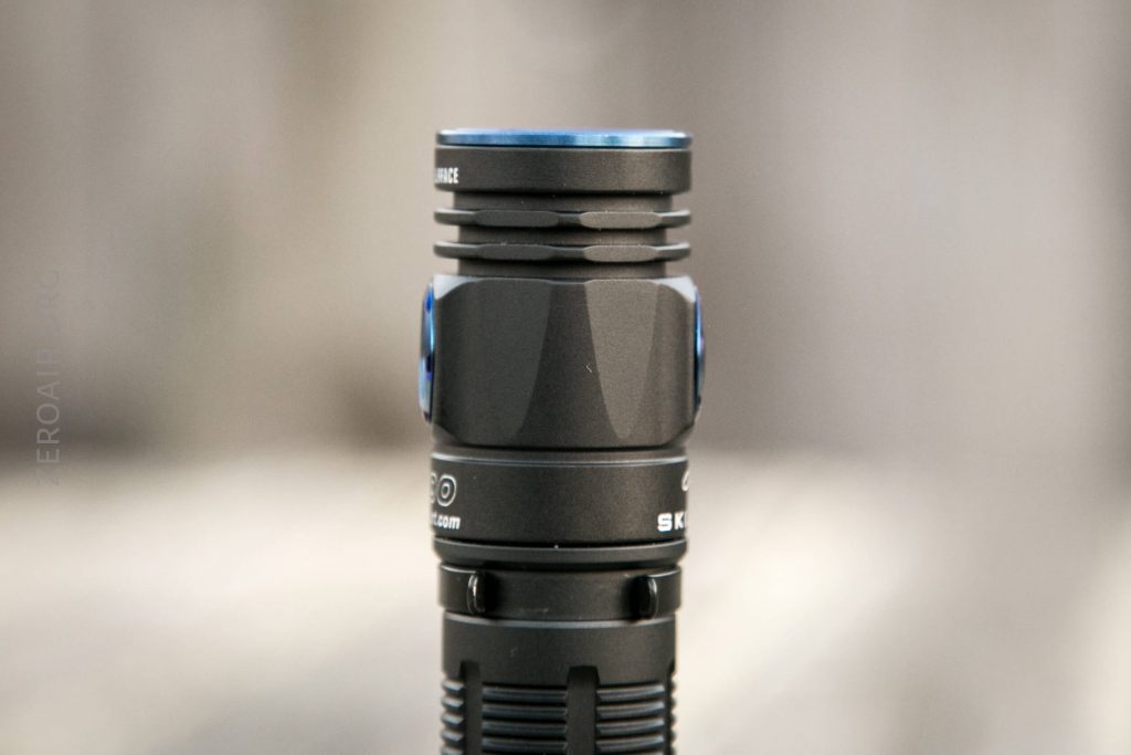 A close-up photo of a black flashlight with blue accents, shown vertically with a blurred neutral background. The flashlight has textured grip details and cooling fins near the head.