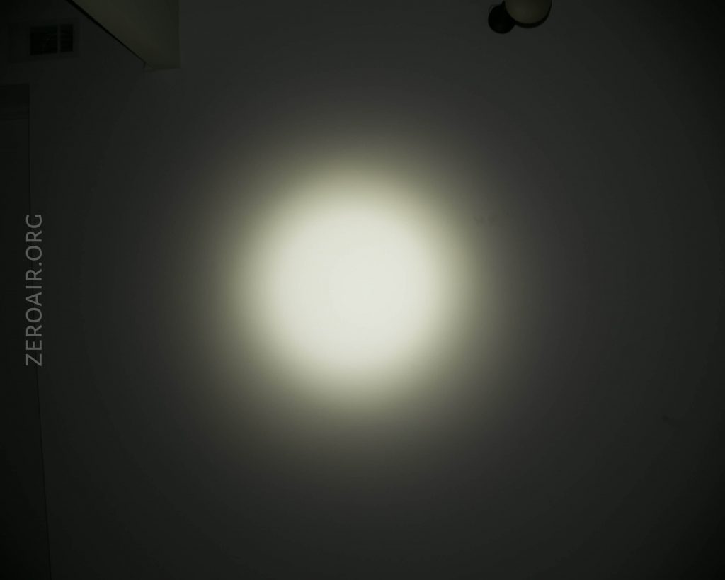 A circular beam of white light is projected onto a dark wall. The text ZEROAIR.ORG is visible vertically on the left side. The edges of the light beam fade gradually into the darkness.