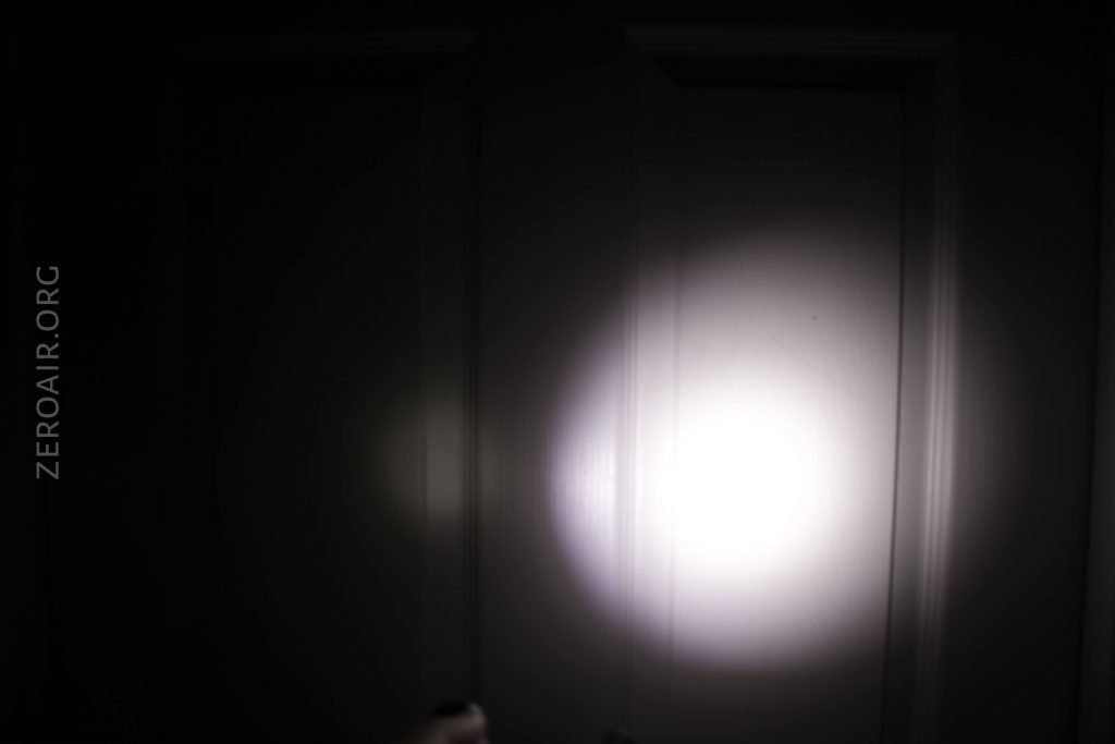 A round, bright white flashlight beam shines on a dark, flat surface, possibly a wall or door. The word ZEROAIR.ORG appears vertically along the left side of the image.