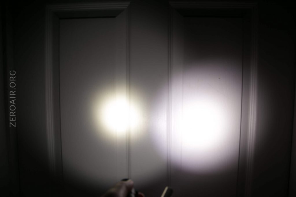 Two flashlight beams, one warm white and one cool white, shine side by side on a light-colored paneled door in a dark environment. A hand holding the flashlights is partially visible at the bottom.